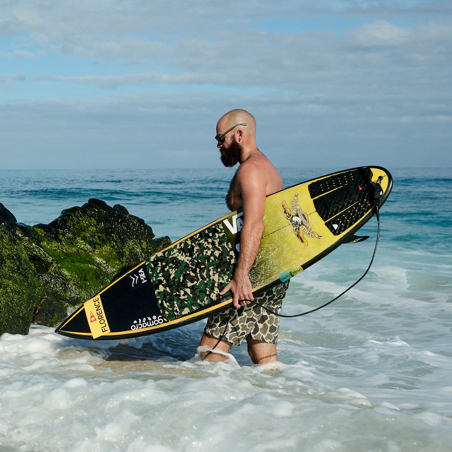 Man holding a surfboard in the ocean with rocks and blue sky in the background
