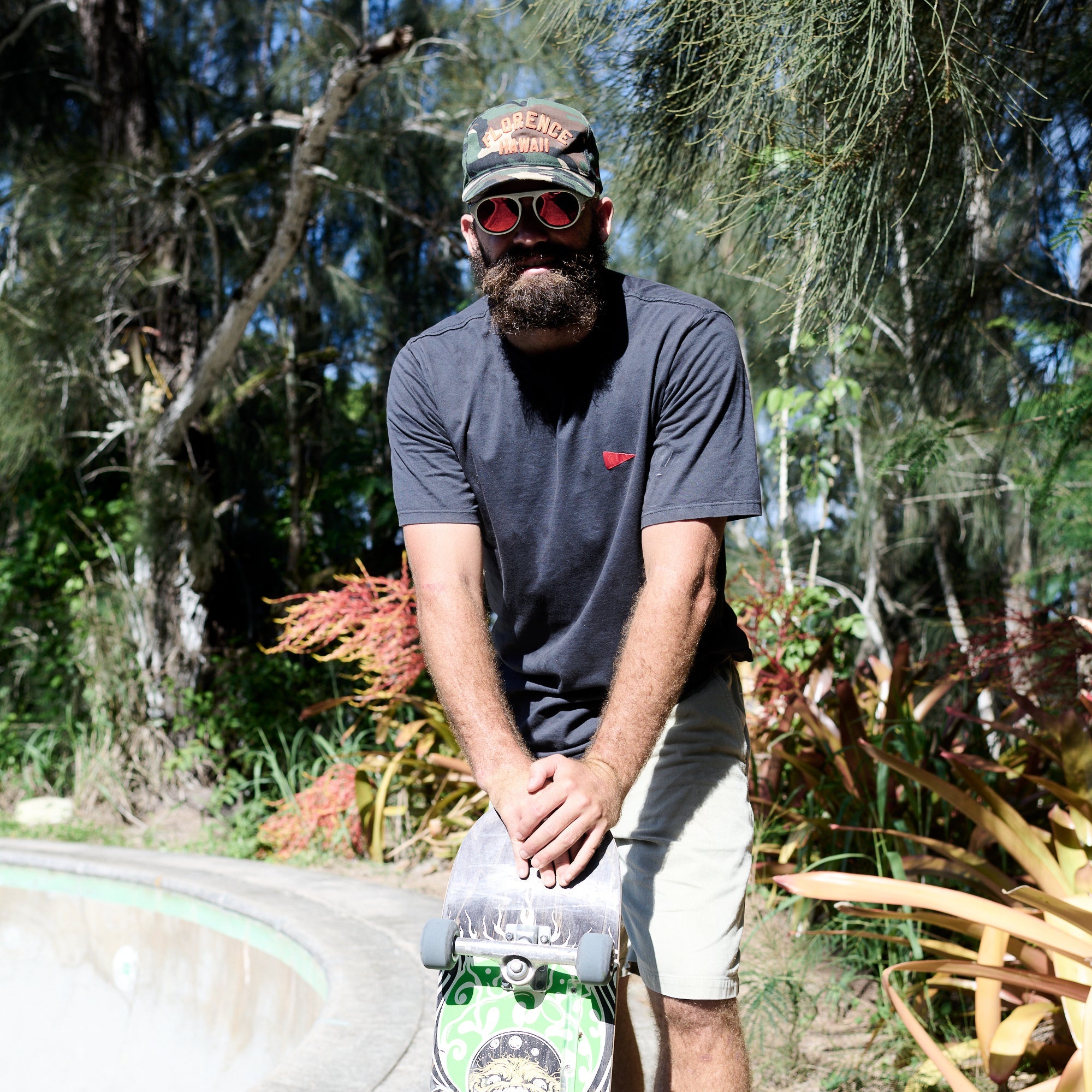 Man with a beard wearing sunglasses, a cap, and a gray t-shirt holding a skateboard in a natural setting.