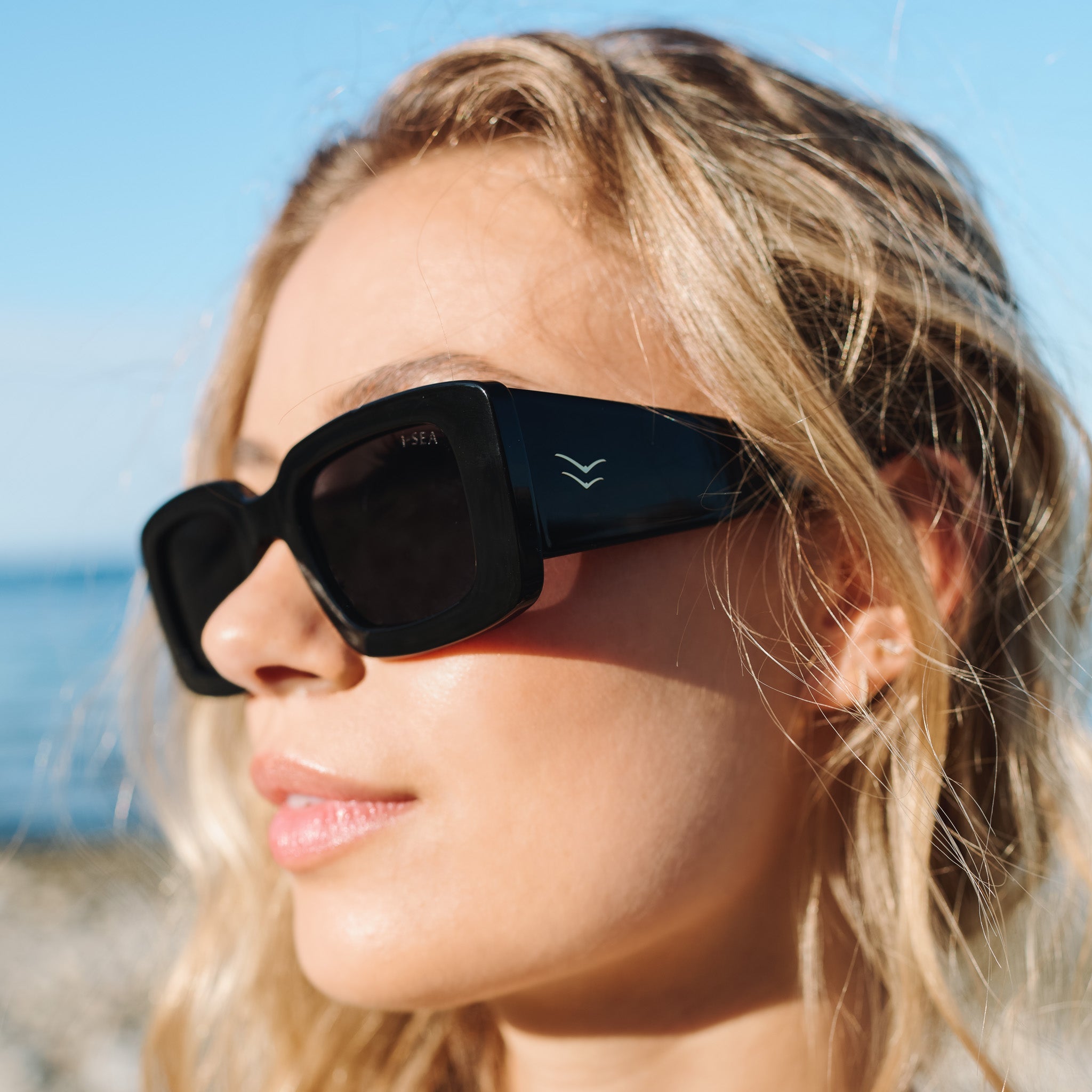 Close-up of a person wearing black sunglasses with a blurred beach background.
BLACK / SMOKE POLARIZED LENS