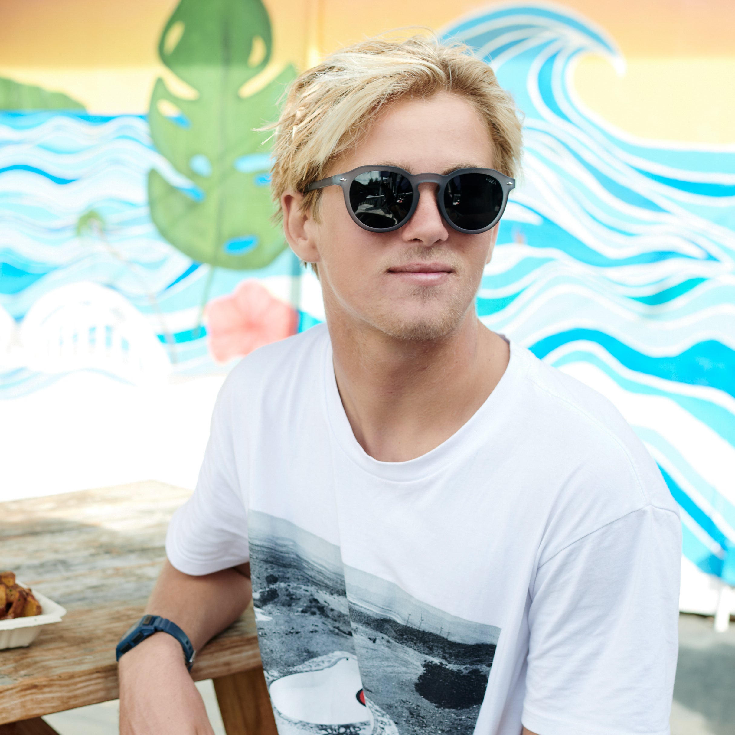 Man wearing sunglasses and a white t-shirt with a graphic design, sitting in front of a colorful mural.
GREY / SMOKE POLARIZED LENS