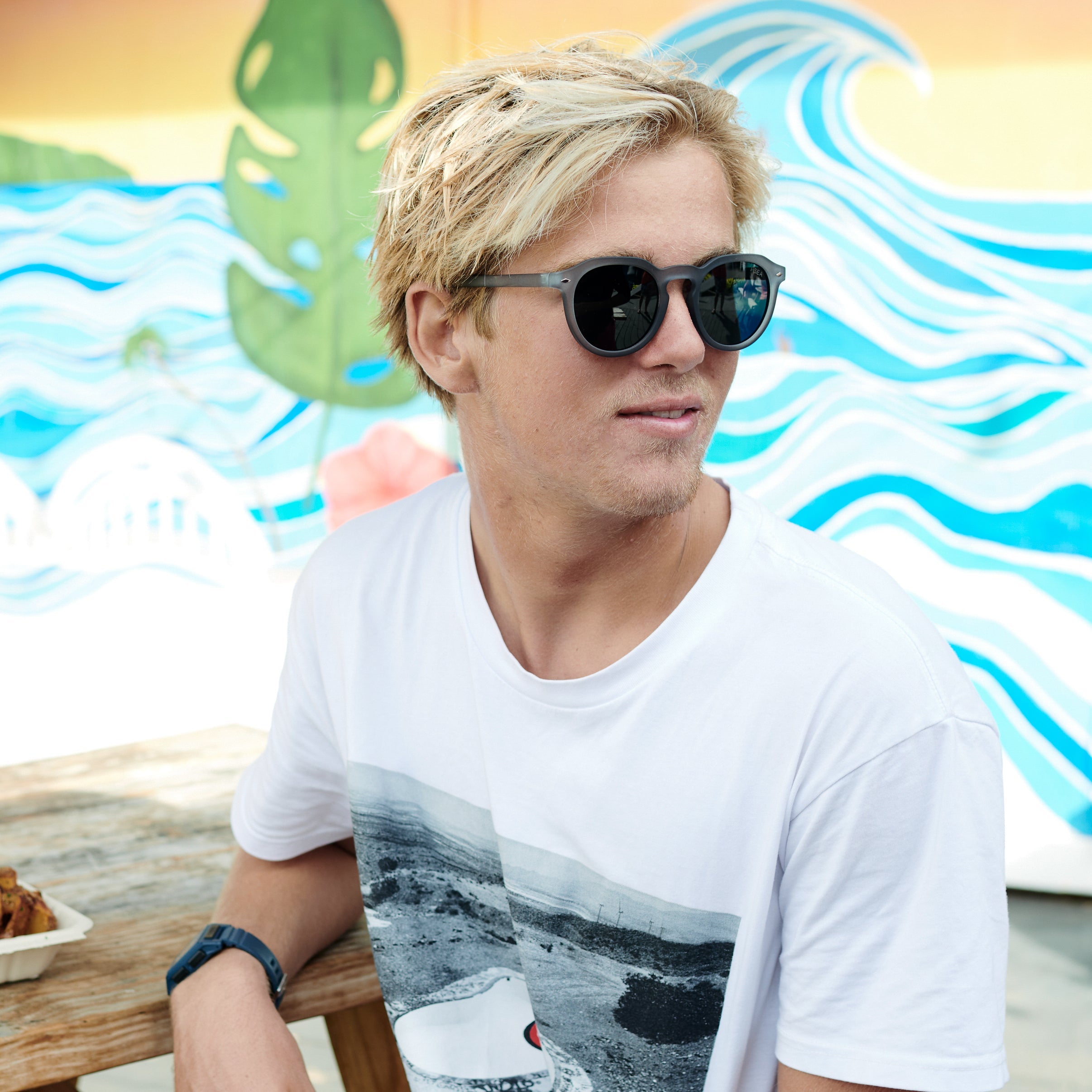 Man wearing sunglasses and a white t-shirt with a graphic design, sitting in front of a colorful mural.
GREY / SMOKE POLARIZED LENS