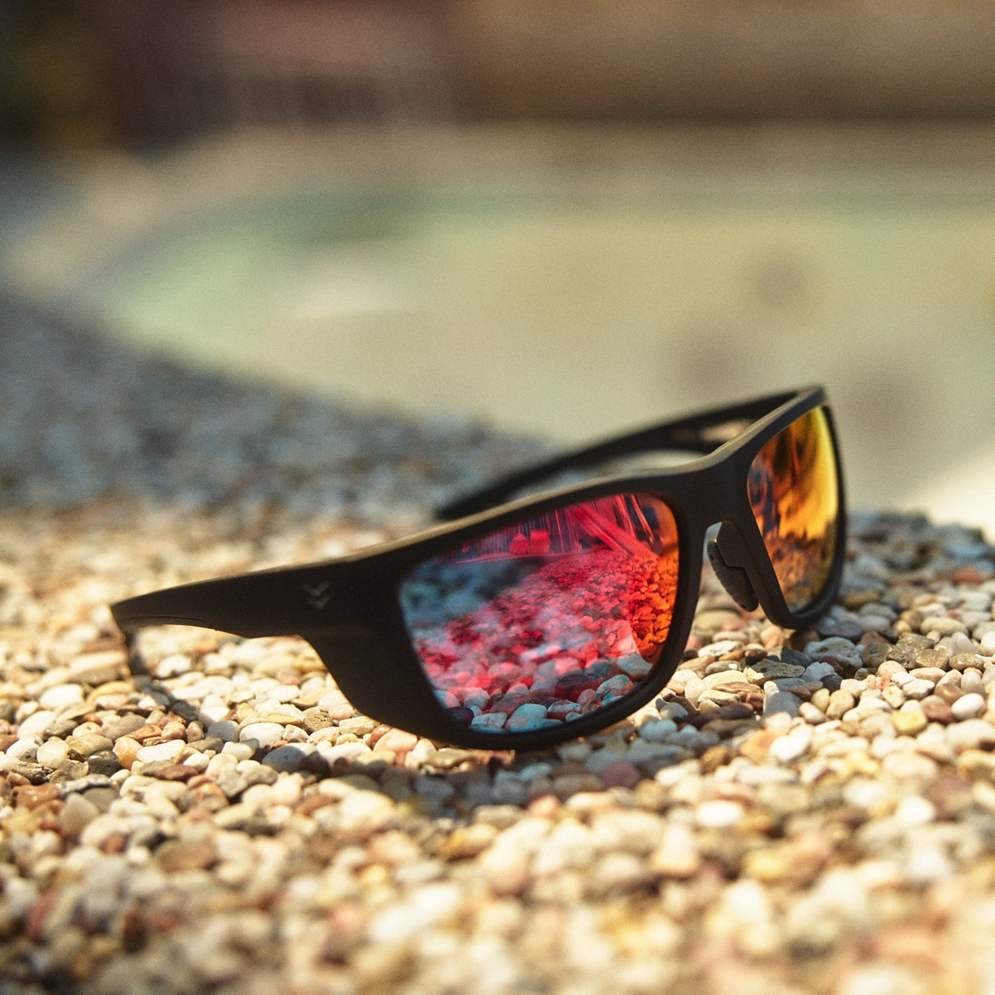 Sunglasses with reflective lenses on a pebbly surface.
BLACK RUBBER / RED MIRROR POLARIZED LENS
