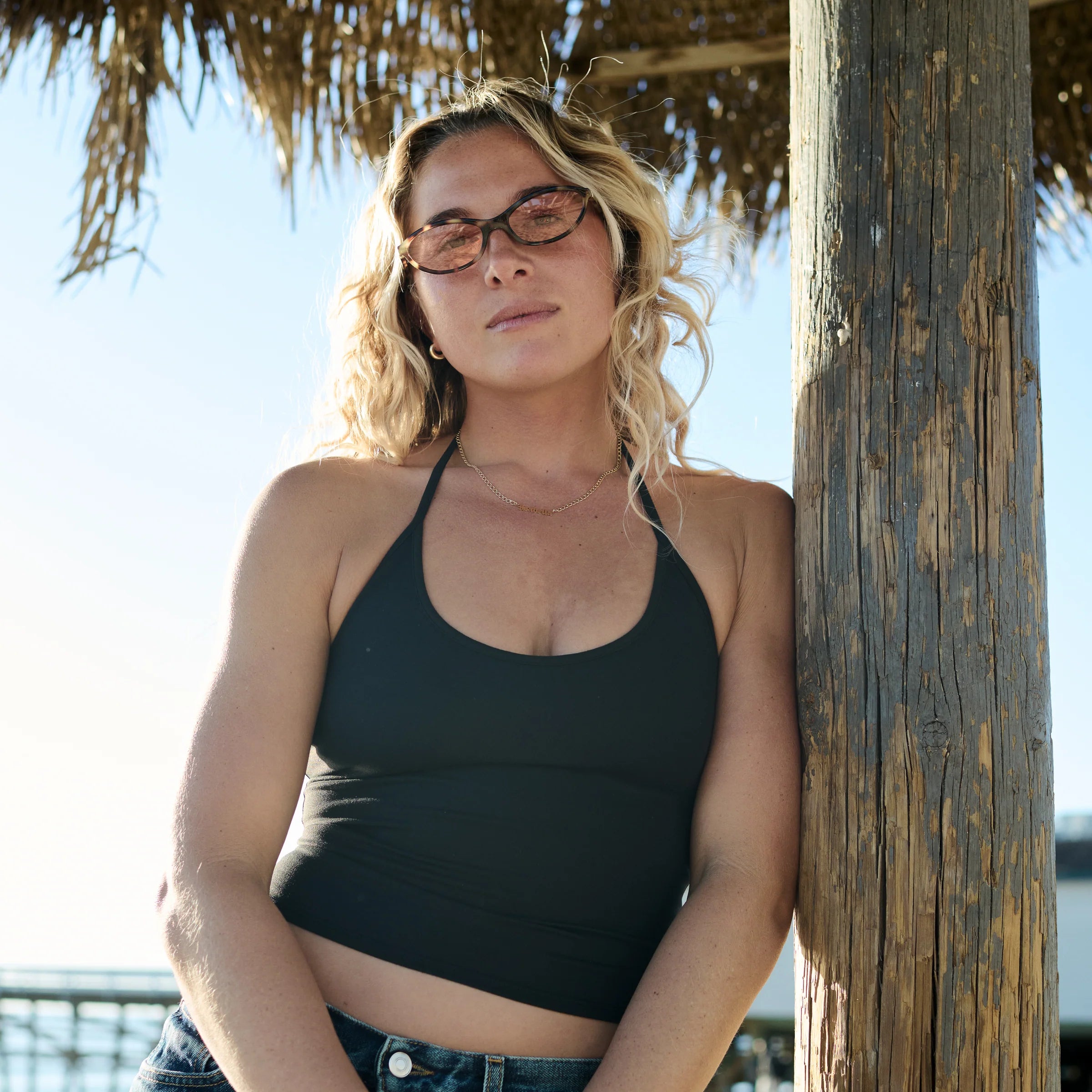 Woman wearing a black tank top and glasses leaning against a wooden post with a beach umbrella in the background.
TAHITI TORT / PEACH LENS