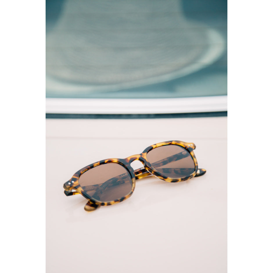 Tortoiseshell sunglasses on a light surface with a blurred background.
TORT / BROWN POLARIZED LENS