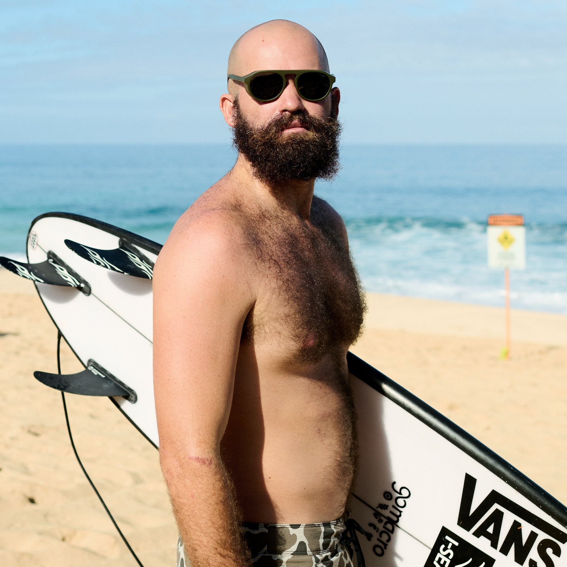 Man with a beard and sunglasses holding a surfboard on a beach