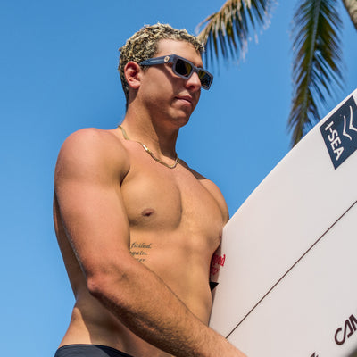 Man holding a surfboard with palm trees and blue sky in the background.
GREY / SMOKE POLARIZED LENS