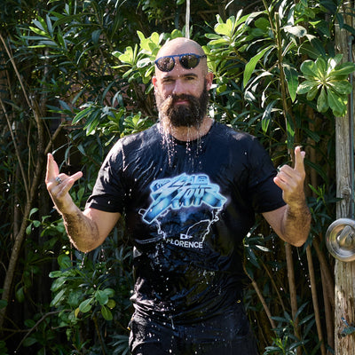Man with sunglasses on head, wet from shower, making hand signs outdoors with greenery.