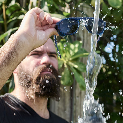Man holding a pair of sunglasses under running water with greenery in the background