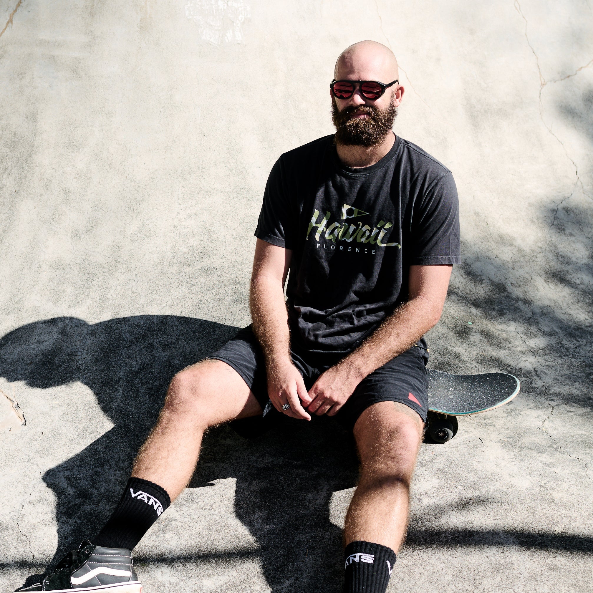 Man sitting on a skateboard wearing a black t-shirt with visible branding, sunglasses, and black shorts.