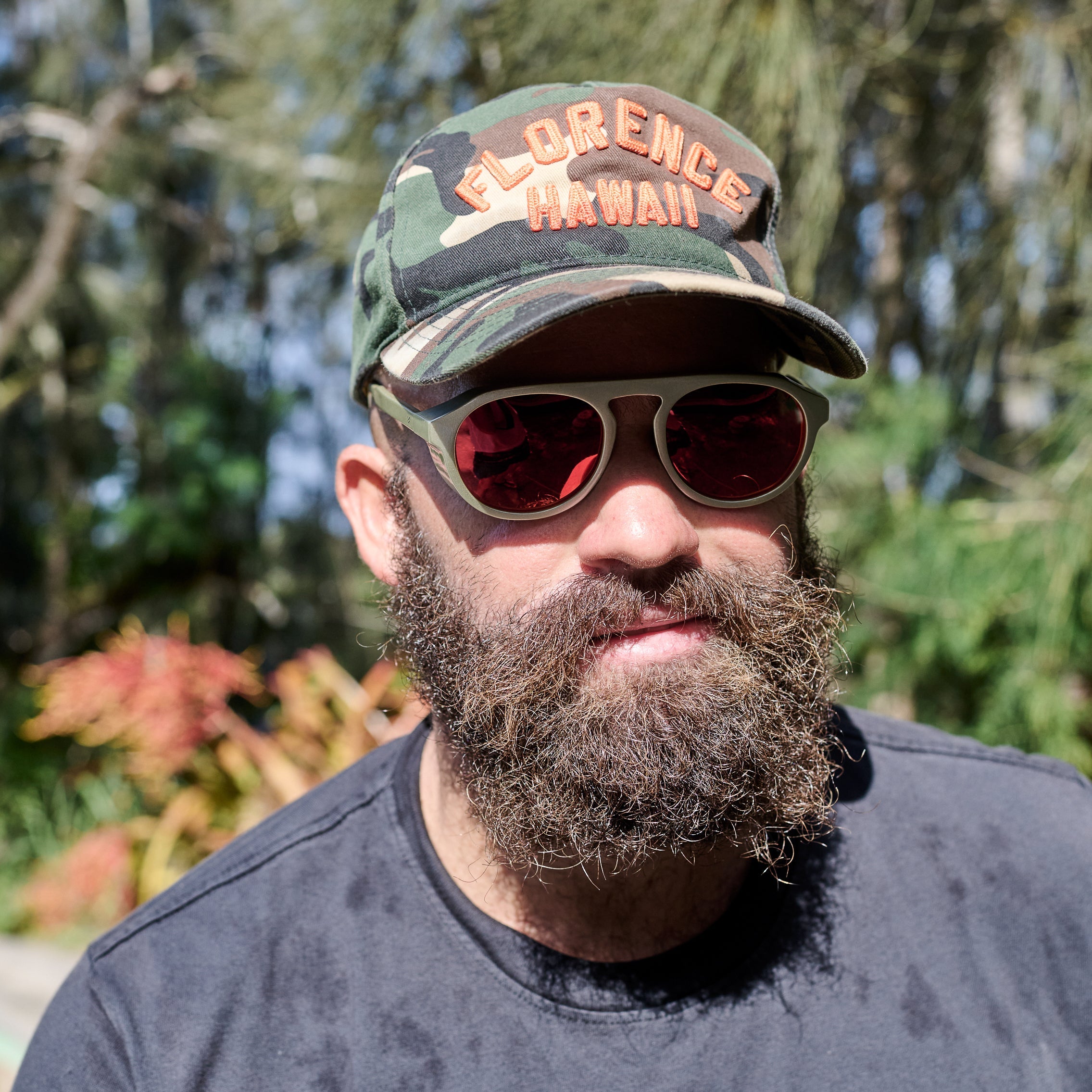 Man wearing a camouflage cap with 'Florence Hawaii' text and sunglasses, standing outdoors with greenery in the background.