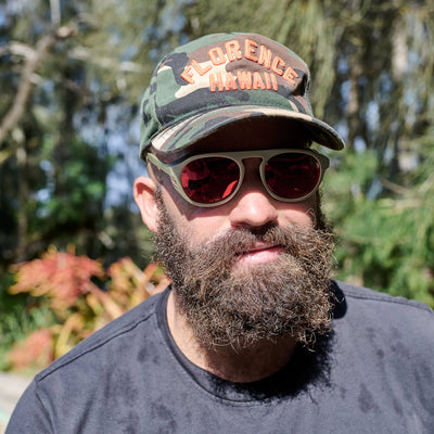 Man wearing a camouflage cap with 'Florence Hawaii' text and sunglasses, standing outdoors with greenery in the background.