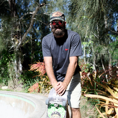 Man with a beard wearing sunglasses, a cap, and a gray t-shirt holding a skateboard in a natural setting.