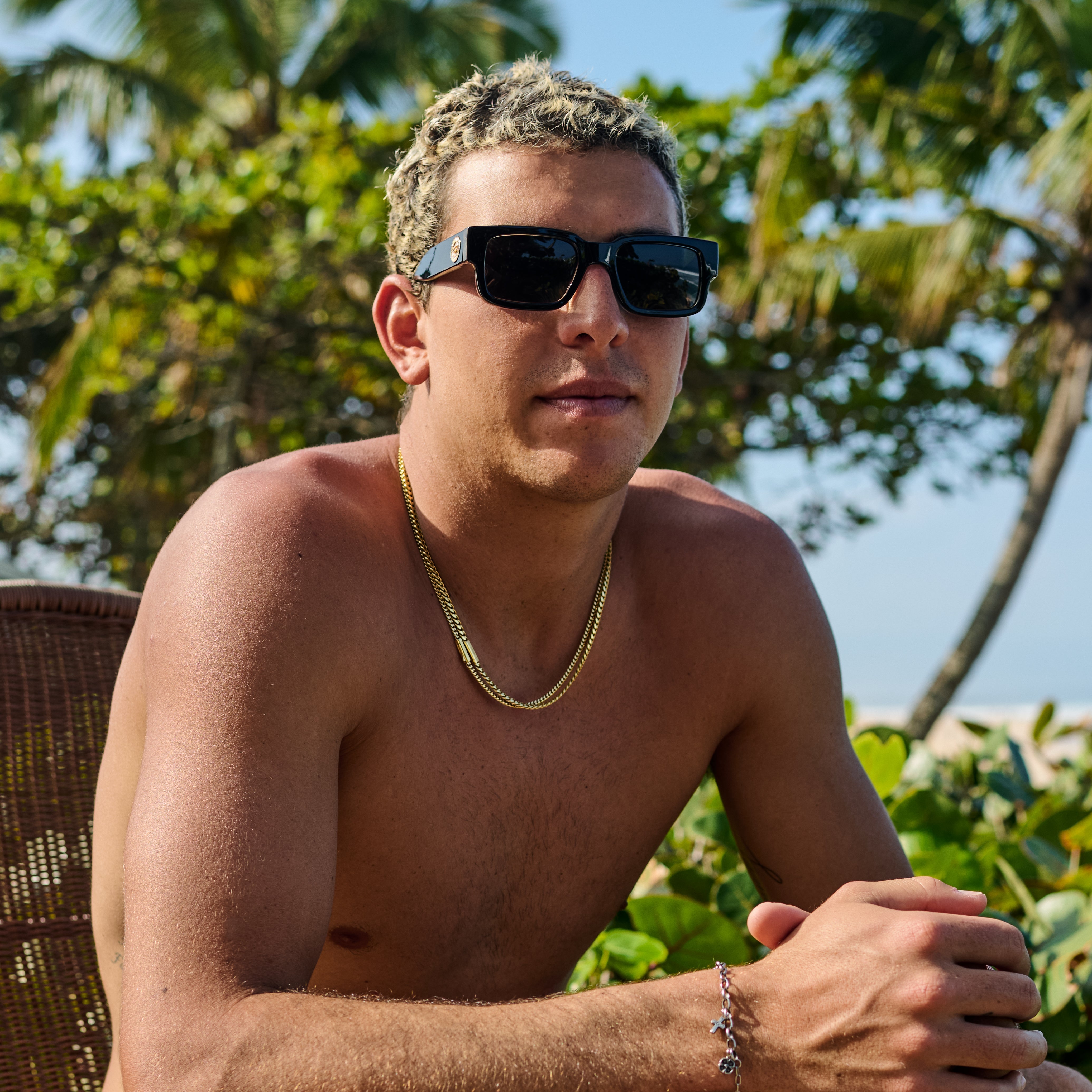 Man wearing sunglasses and a gold chain, sitting outdoors with greenery in the background.
BLACK LEOPARD / SMOKE POLARIZED LENS