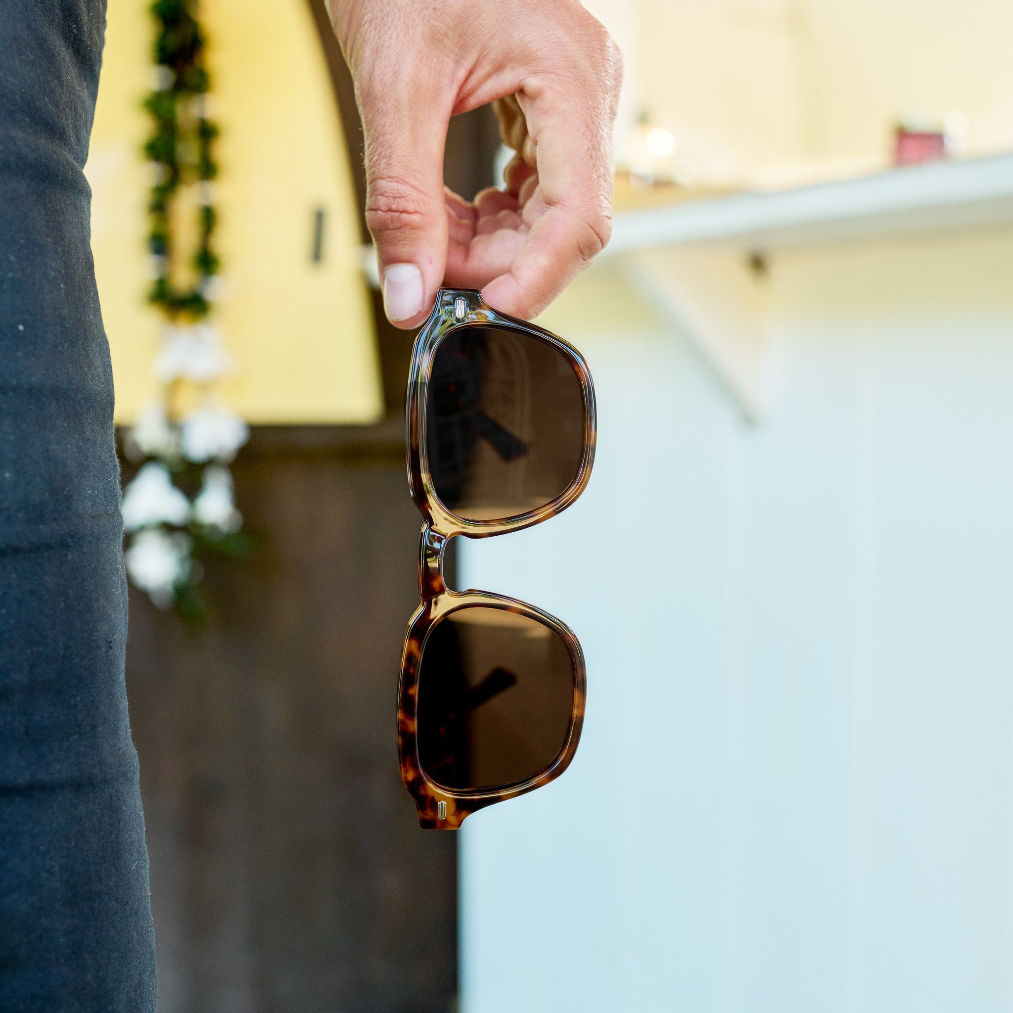 Person holding a pair of sunglasses with a blurred background.
TORT / BROWN POLARIZED LENS