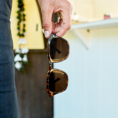 Person holding a pair of sunglasses with a blurred background.
TORT / BROWN POLARIZED LENS