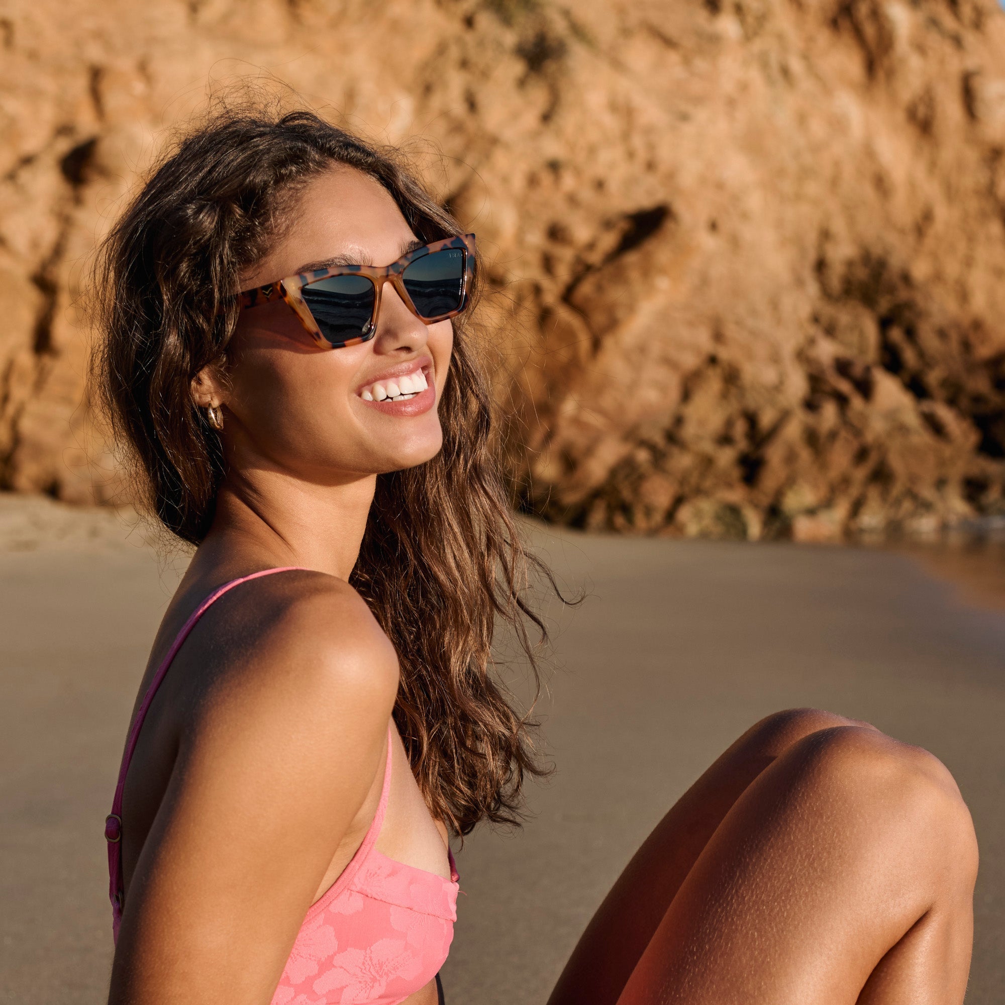 Woman in a pink bikini and sunglasses sitting on a beach with rocky background.
TORT / SMOKE POLARIZED LENS