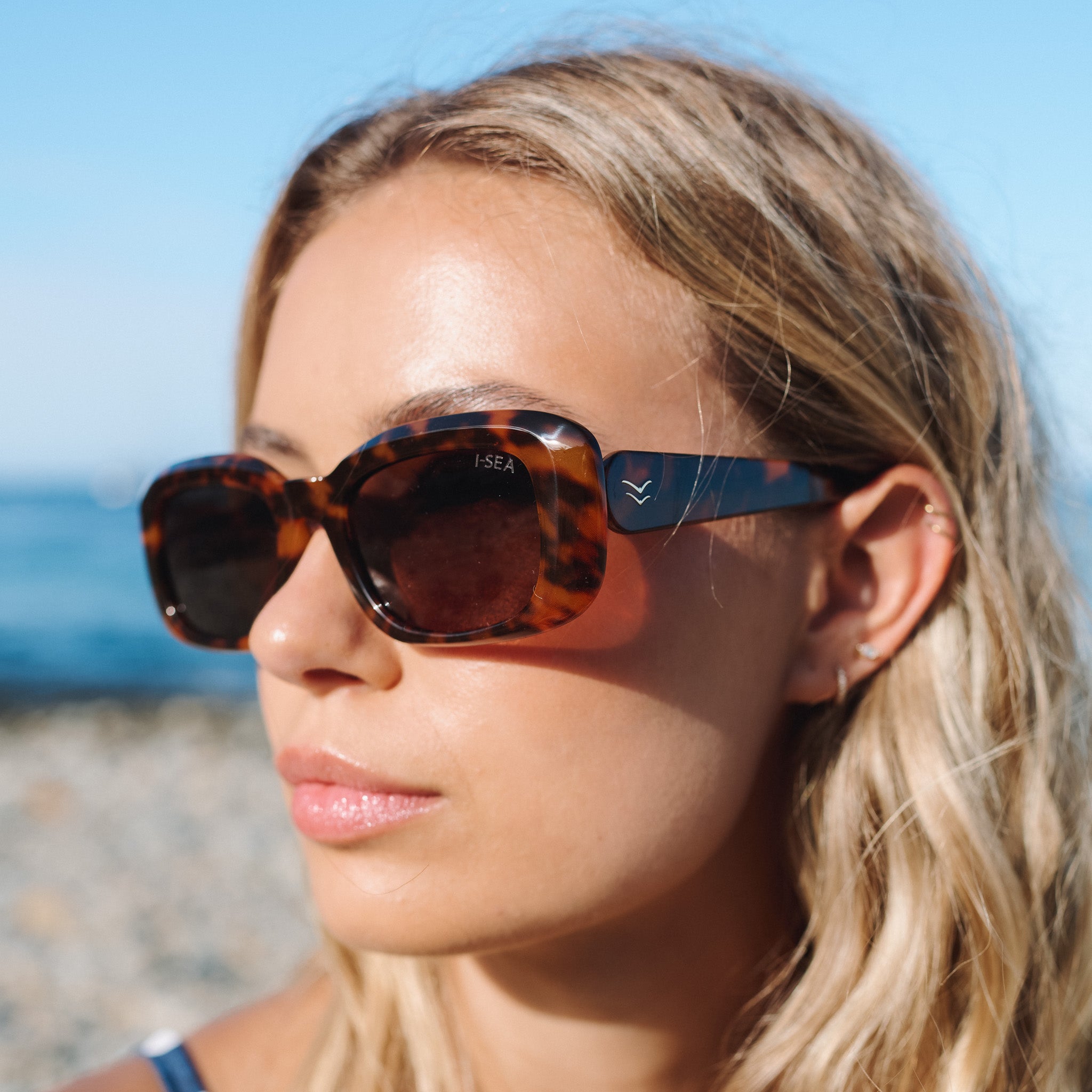 Woman wearing tortoiseshell sunglasses with a blurred beach background.
TORT / BROWN POLARIZED LENS