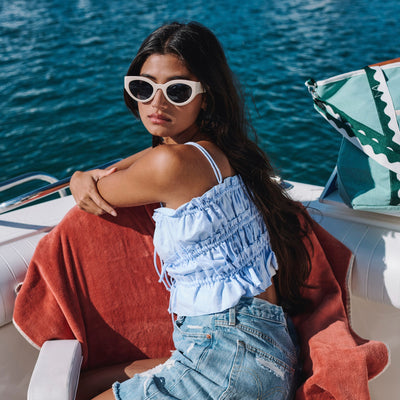 Woman sitting on a boat wearing sunglasses and a blue outfit with a red towel.
CREAM / NAVY POLARIZED LENS