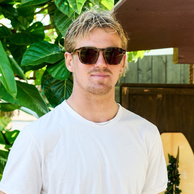 Man wearing sunglasses and a white t-shirt outdoors with greenery in the background.
TORT / BROWN POLARIZED LENS