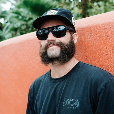 Man with a beard wearing sunglasses and a cap, standing next to an orange wall with greenery in the background.
INK / SMOKE POLARIZED LENS