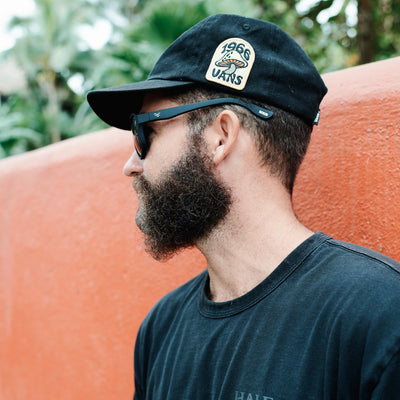 Man wearing a black cap with a Vans logo, sunglasses, and a dark t-shirt against an orange wall with greenery.
INK / SMOKE POLARIZED LENS