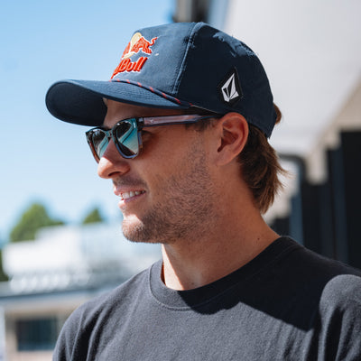 Man wearing a navy cap with a logo and sunglasses, outdoors.
TORT COLA / BROWN POLARIZED LENS