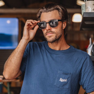 Man wearing sunglasses and a blue t-shirt with a logo in an indoor setting.
BLACK / SMOKE POLARIZED LENS