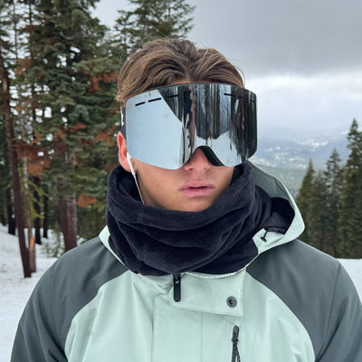 Person wearing reflective goggles and a black scarf in a snowy landscape with trees.
OLIVE / SILVER
