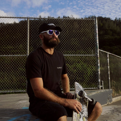 Man with sunglasses and a beard sitting on a bench holding a skateboard, with a chain-link fence and trees in the background.