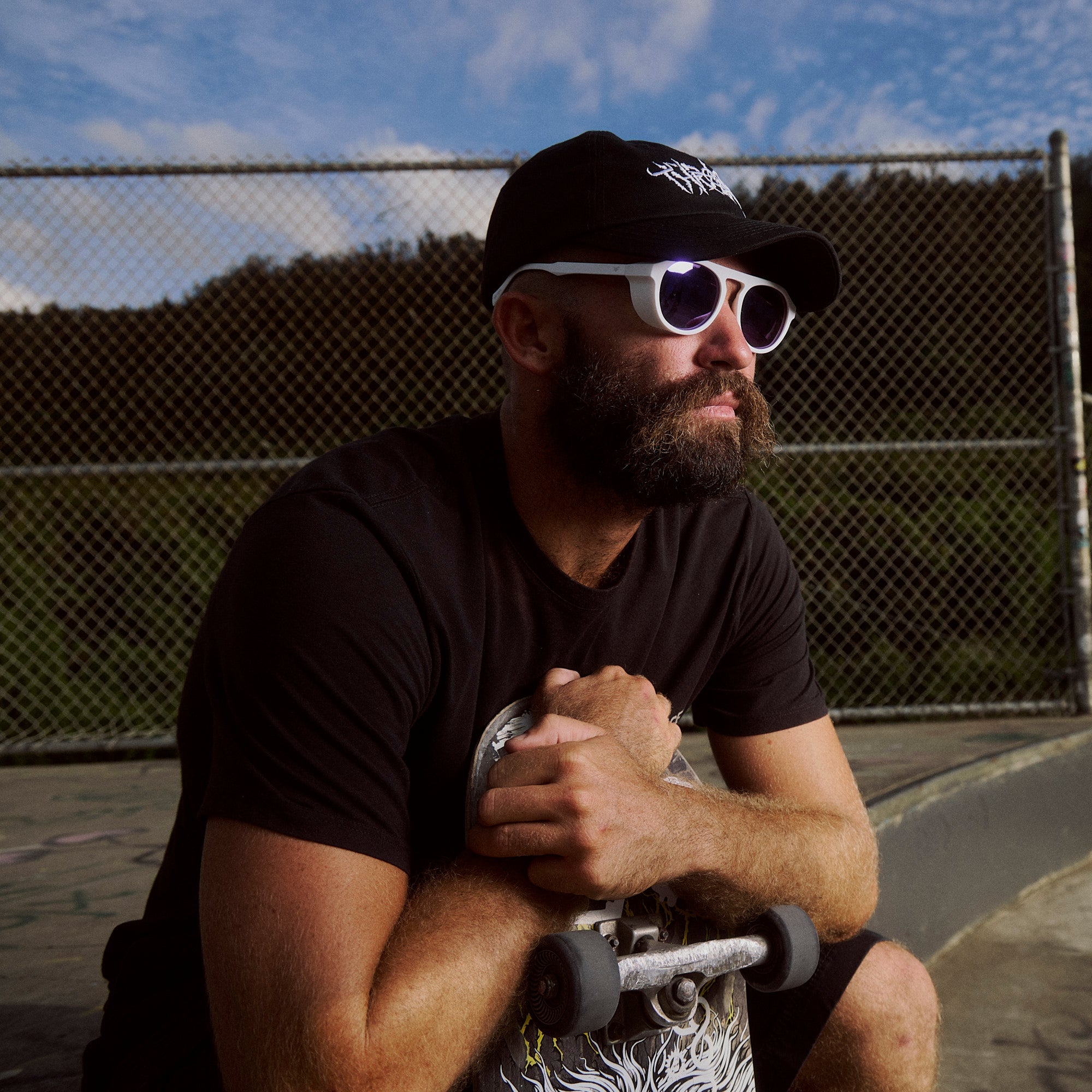 Man with a beard wearing sunglasses and a black cap, sitting with a skateboard at a skate park.