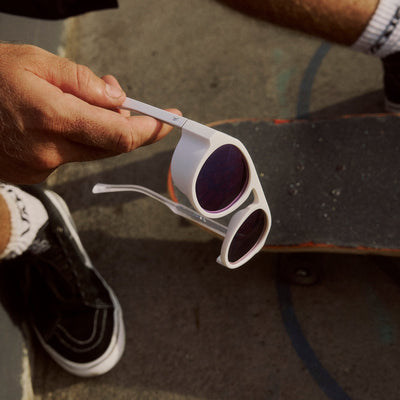 Person holding white sunglasses with purple lenses on a skateboard