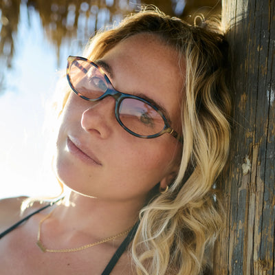 Woman wearing glasses leaning against a wooden wall with sunlight filtering through.
TAHITI TORT / PEACH LENS