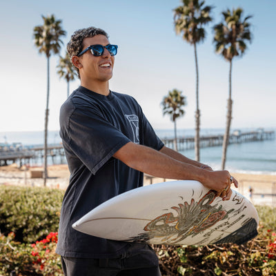 Man holding a surfboard with palm trees and a pier in the background.
DARK SEAS / SMOKE POLARIZED LENS