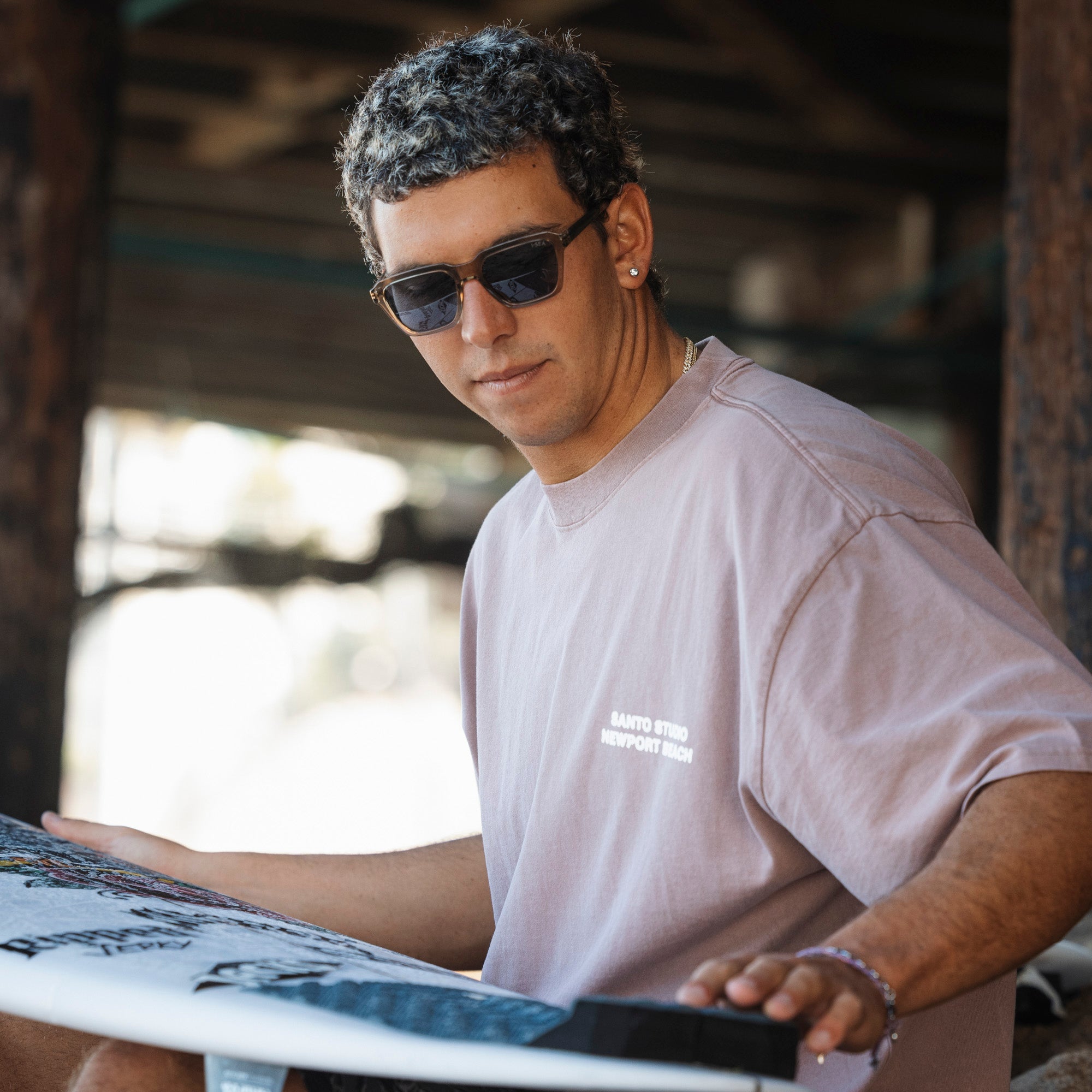 Man wearing sunglasses and a light-colored t-shirt holding a surfboard under a wooden shelter.
TAUPE / NAVY POLARIZED LENS