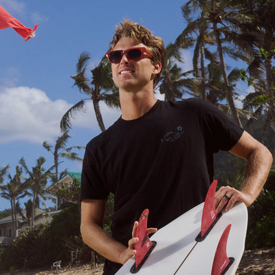 Man holding a surfboard with palm trees and a clear sky in the background.
TANGERINE / NAVY POLARIZED LENS