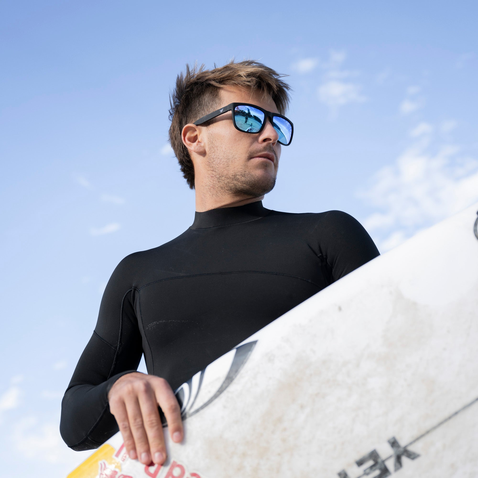 Man in a black wetsuit holding a surfboard with a blue sky background.
BLACK / ICE BLUE POLARIZED LENS