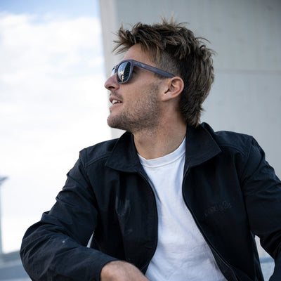 Man wearing sunglasses and a black jacket over a white shirt, sitting outdoors.
GREY / SMOKE POLARIZED LENS