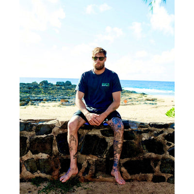 Man sitting on a stone wall by the beach wearing sunglasses and a blue t-shirt.
TORT / BROWN POLARIZED LENS