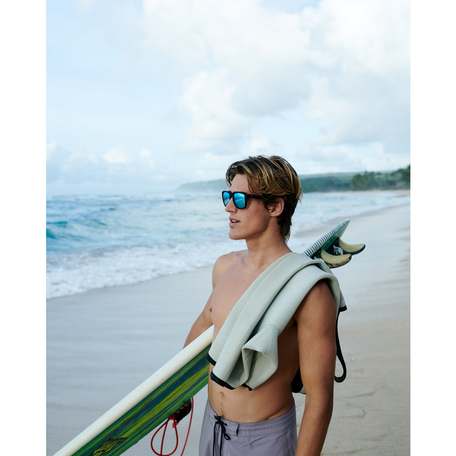 Man with a towel draped over his shoulder holding a surfboard on a beach.
BLACK / SILVER BLUE POLARIZED LENS