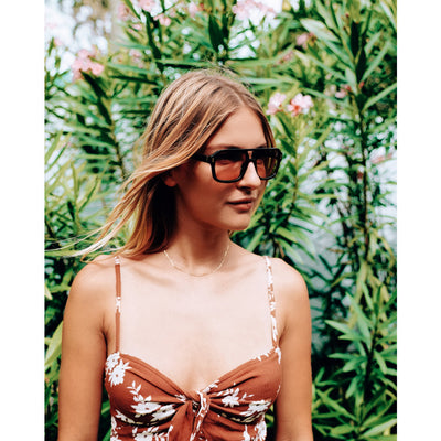 Woman wearing sunglasses and a floral dress in front of green foliage.
BLACK / PEACH LENS